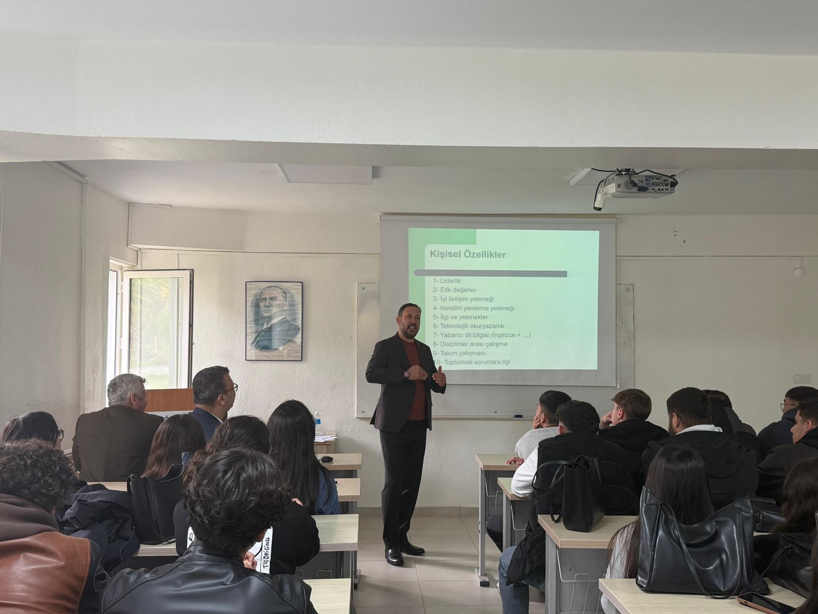 A presentation taking place in a classroom at Dalaman Vocational School. A speaker stands at the front, explaining a slide titled ‘Personal Qualities.’ Students seated at their desks are following the presentation. An Atatürk portrait and large windows are visible on the walls.
